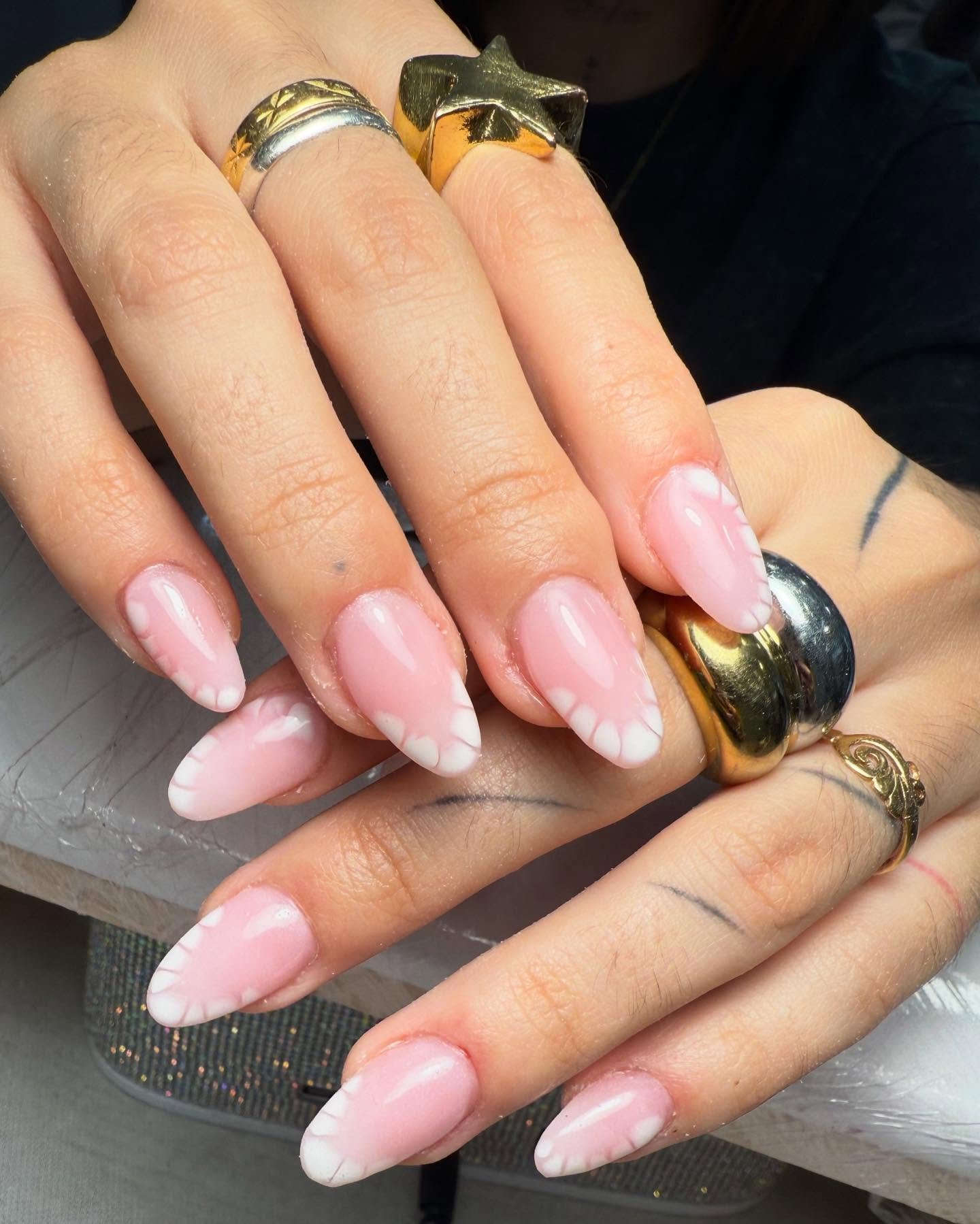 Manos con uñas largas en forma almendrada, esmaltadas en tono rosa claro con diseño de líneas blancas onduladas en las puntas. La persona lleva varios anillos dorados y plateados.