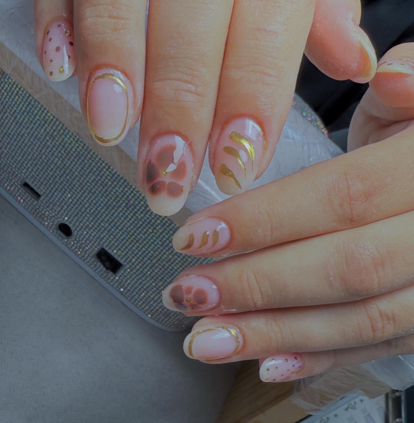 Manos con uñas esmaltadas en tonos nude y decoraciones variadas. Algunas uñas tienen flores marrones, otras detalles dorados y puntos rojos sobre fondo blanco.