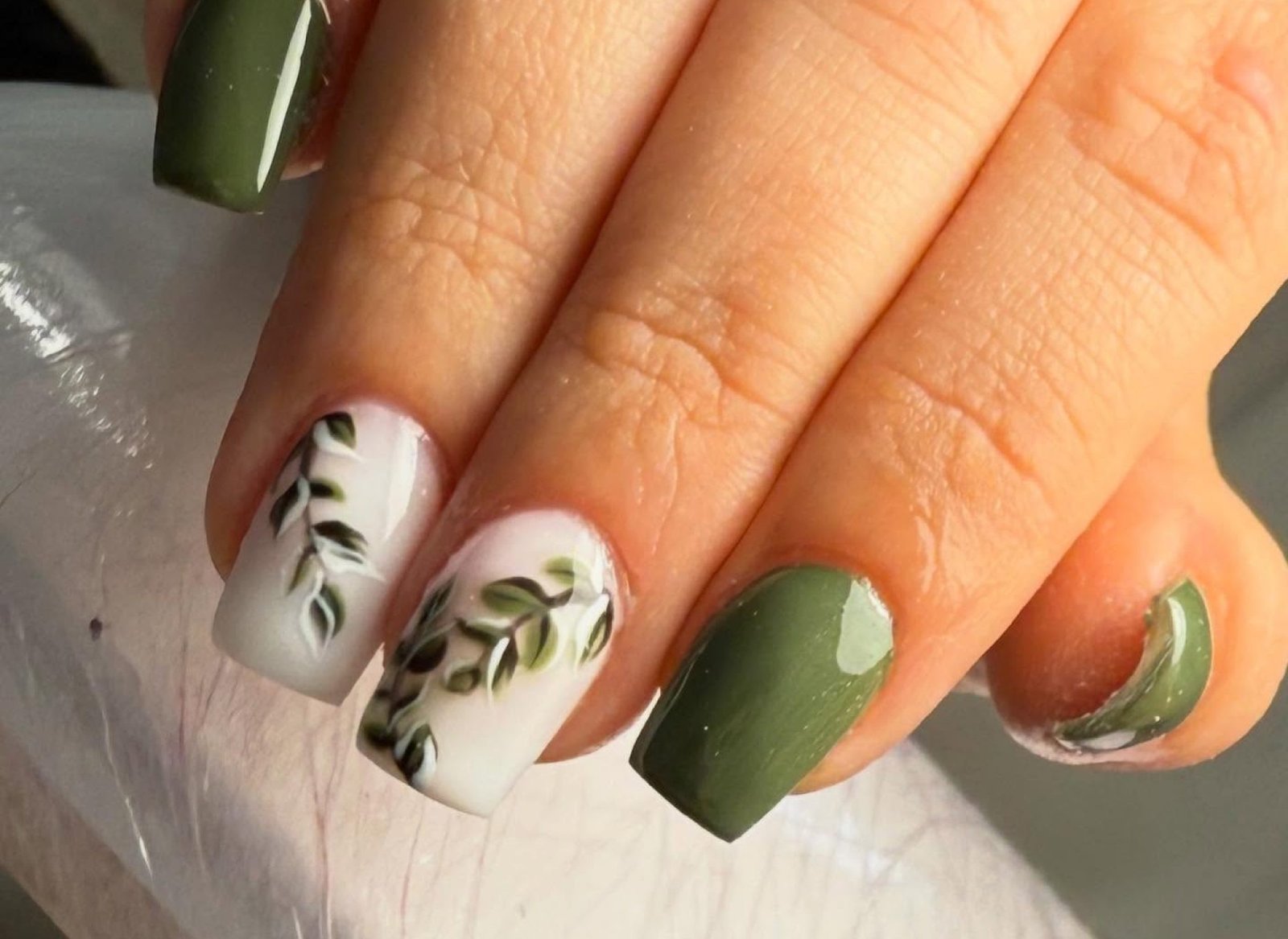 Manos con uñas cortas esmaltadas en verde oscuro y blanco. Dos uñas blancas tienen diseño de ramas con hojas verdes.
