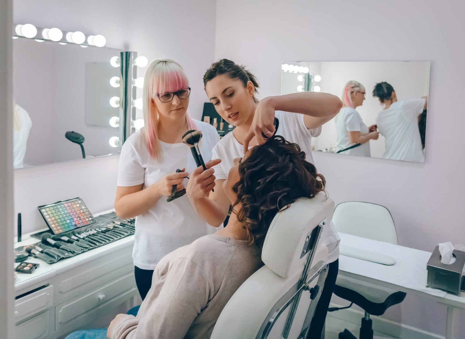 Dos esteticistas maquillando a una clienta sentada en una silla blanca frente a un tocador iluminado. Una sostiene una brocha y la otra un delineador.